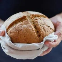 Dłonie trzymają okrągły bochenek chleba, owinięty w lnianą ściereczkę. Panie dobry jak chleb, symbolizuje świeżość i tradycję.