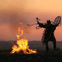 Sylwetka w tradycyjnym stroju, z bębnem, tańczy przy ognisku. Scena oddaje klimat szamanizmu podczas zachodu słońca.