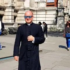 Uśmiechnięty pastor w czarnej sutannie i okularach przeciwsłonecznych idzie ulicą. W tle widać ludzi i budynek.