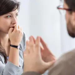 Kobieta z **skrupułami** gryzie paznokcie, słuchając mężczyzny w okularach.