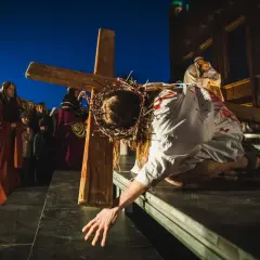 Scena kiedy jest Droga Krzyżowa: Jezus w cierniowej koronie, z krwią na szacie, upada pod ciężarem krzyża, podczas gdy tłum obserwuje.
