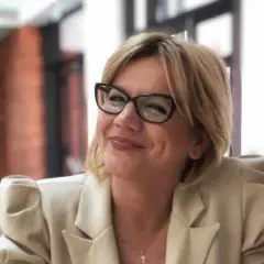 Uśmiechnięta kobieta w okularach i jasnej marynarce, z naszyjnikiem z krzyżykiem. Czy to objawy przekleństwa w rodzinie, czy tylko zmęczenie?