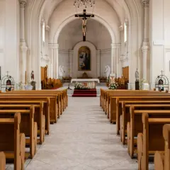 Wnętrze kościoła z rzędami drewnianych ławek. W ołtarzu głównym wisi obraz, a nad nim krucyfiks. 1 maja święto kościelne, czas zadumy.