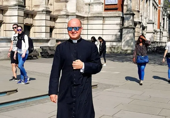 Uśmiechnięty pastor w czarnej sutannie i okularach przeciwsłonecznych idzie ulicą. W tle widać ludzi i budynek.
