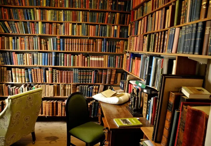 Wnętrze biblioteki z regałami pełnymi książek. Na biurku leży poduszka z książkami, a obok krzesło. Nawet niewierzący doceniłby bogactwo wiedzy.