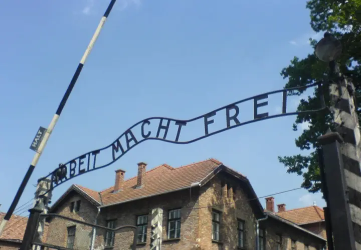 Brama z napisem "Arbeit Macht Frei" nad ceglanymi budynkami pod błękitnym niebem.