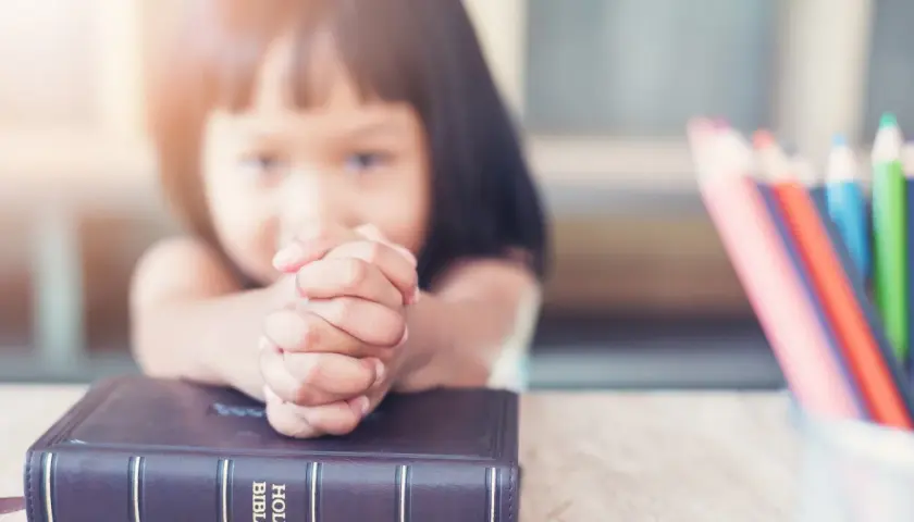 Dziecko z dłońmi złożonymi do modlitwy nad Biblią. Obok kolorowe kredki, symbol nauki dekalogu 10 przykazań.