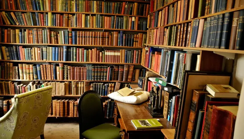 Wnętrze biblioteki z regałami pełnymi książek. Na biurku leży poduszka z książkami, a obok krzesło. Nawet niewierzący doceniłby bogactwo wiedzy.