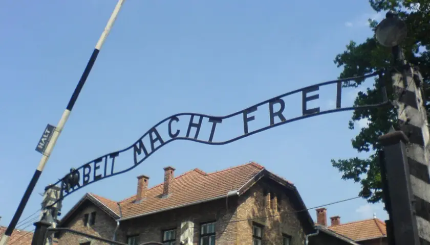 Brama z napisem "Arbeit Macht Frei" nad ceglanymi budynkami pod błękitnym niebem.