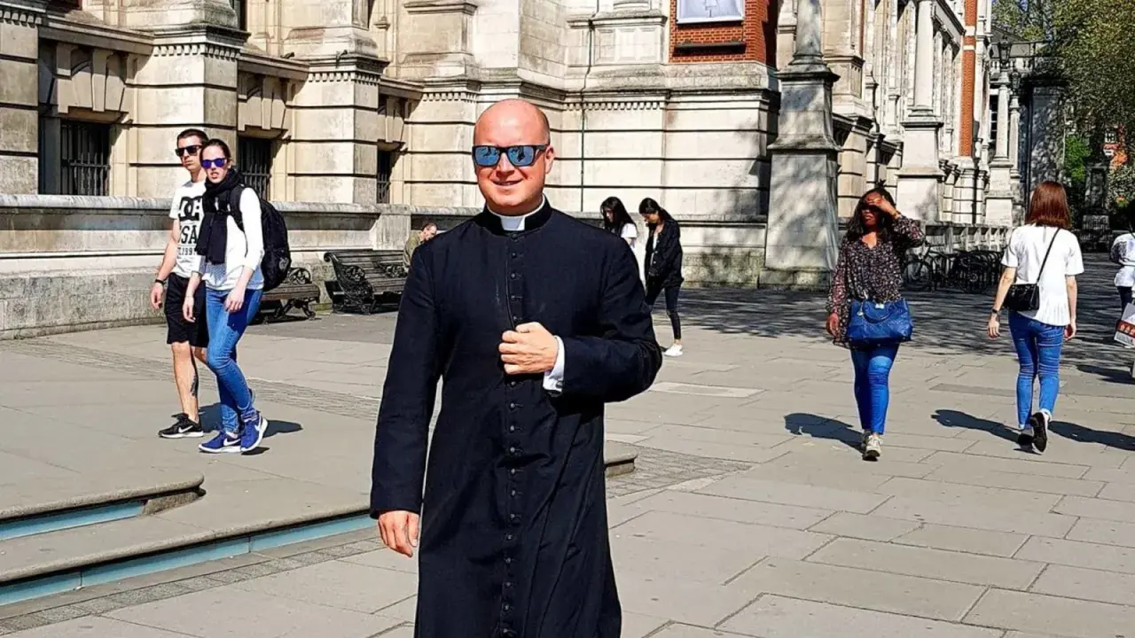 Uśmiechnięty pastor w czarnej sutannie i okularach przeciwsłonecznych idzie ulicą. W tle widać ludzi i budynek.