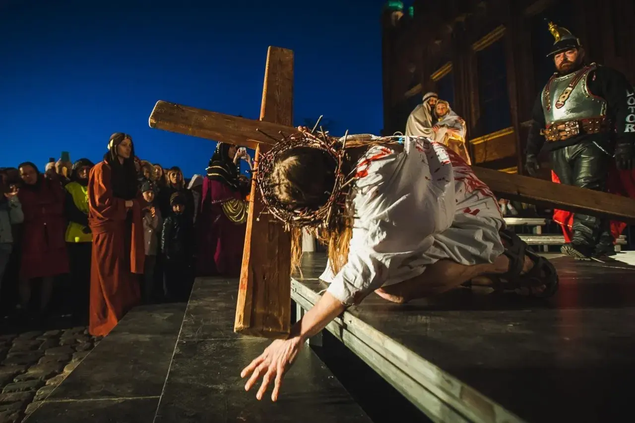 Scena kiedy jest Droga Krzyżowa: Jezus w cierniowej koronie, z krwią na szacie, upada pod ciężarem krzyża, podczas gdy tłum obserwuje.