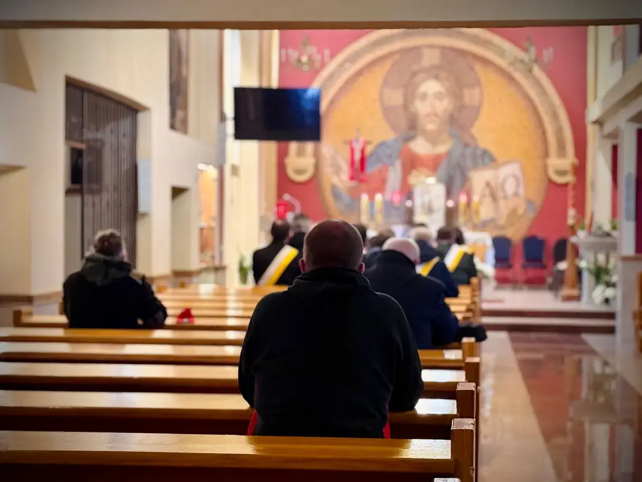 Ludzie siedzą w kościele, pogrążeni w różnych rodzajach modlitwy. W tle widać mozaikę z Jezusem.
