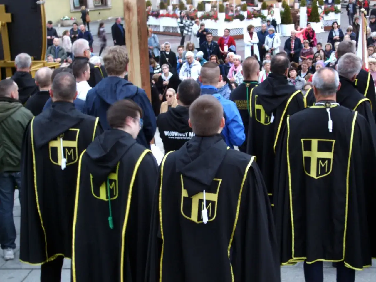 Grupa mężczyzn w czarnych płaszczach z żółtymi obszyciami i symbolem krzyża z literą M. To rycerze Maryi, gotowi do uroczystości.