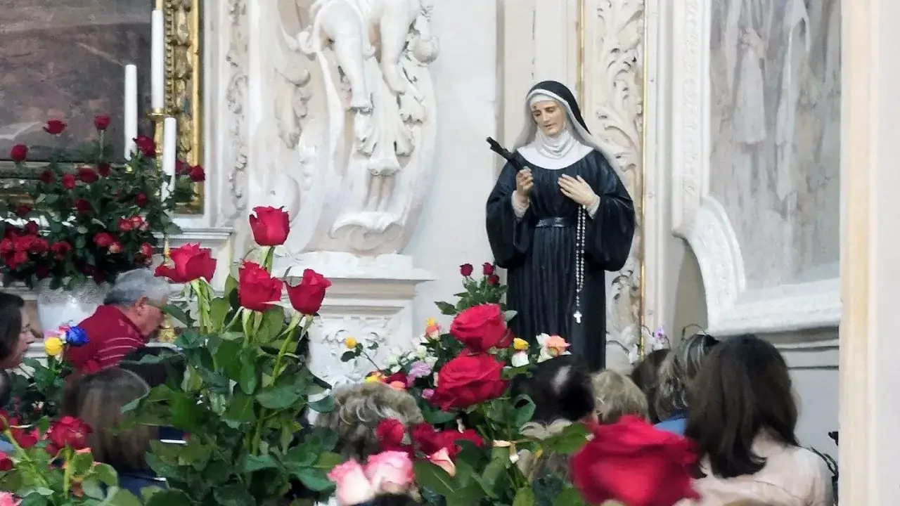 W kościele, przy ołtarzu z figurą świętej, wierni słuchają o przepowiedniach św. Rity. Wokół mnóstwo czerwonych róż.
