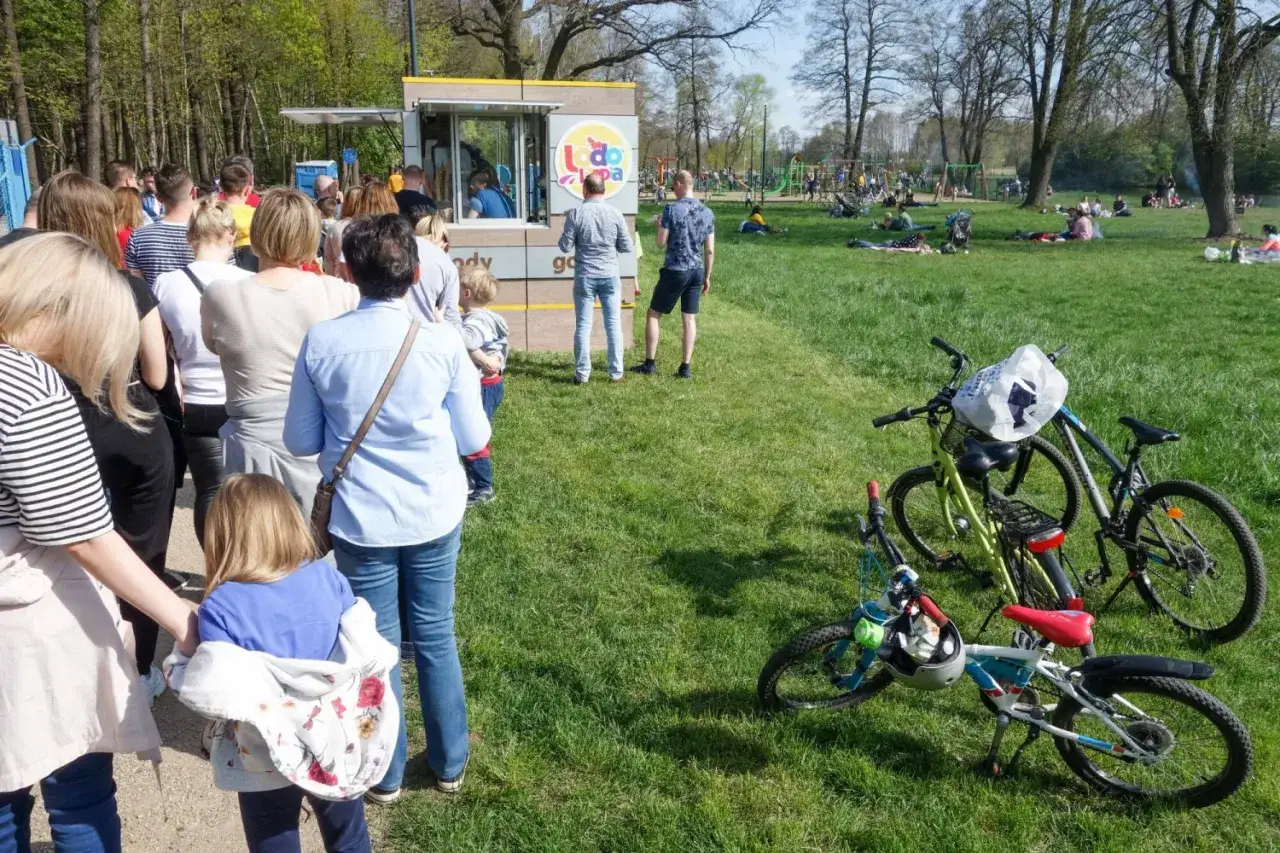 Ludzie w parku, kolejka do budki z lodami. Czy 3 maja można pracować? Widać rowery, plac zabaw i piknikujących.