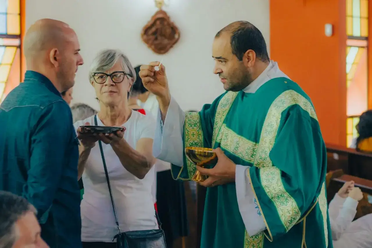 Pastor w zielonej szacie udziela komunii świętej. Kobieta z okularami i mężczyzna w dżinsowej koszuli przyjmują hostię.