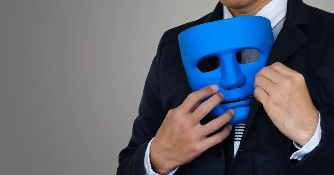 Mężczyzna w garniturze trzyma niebieską maskę, symbolizującą hipokrytę, który ukrywa swoje prawdziwe oblicze.